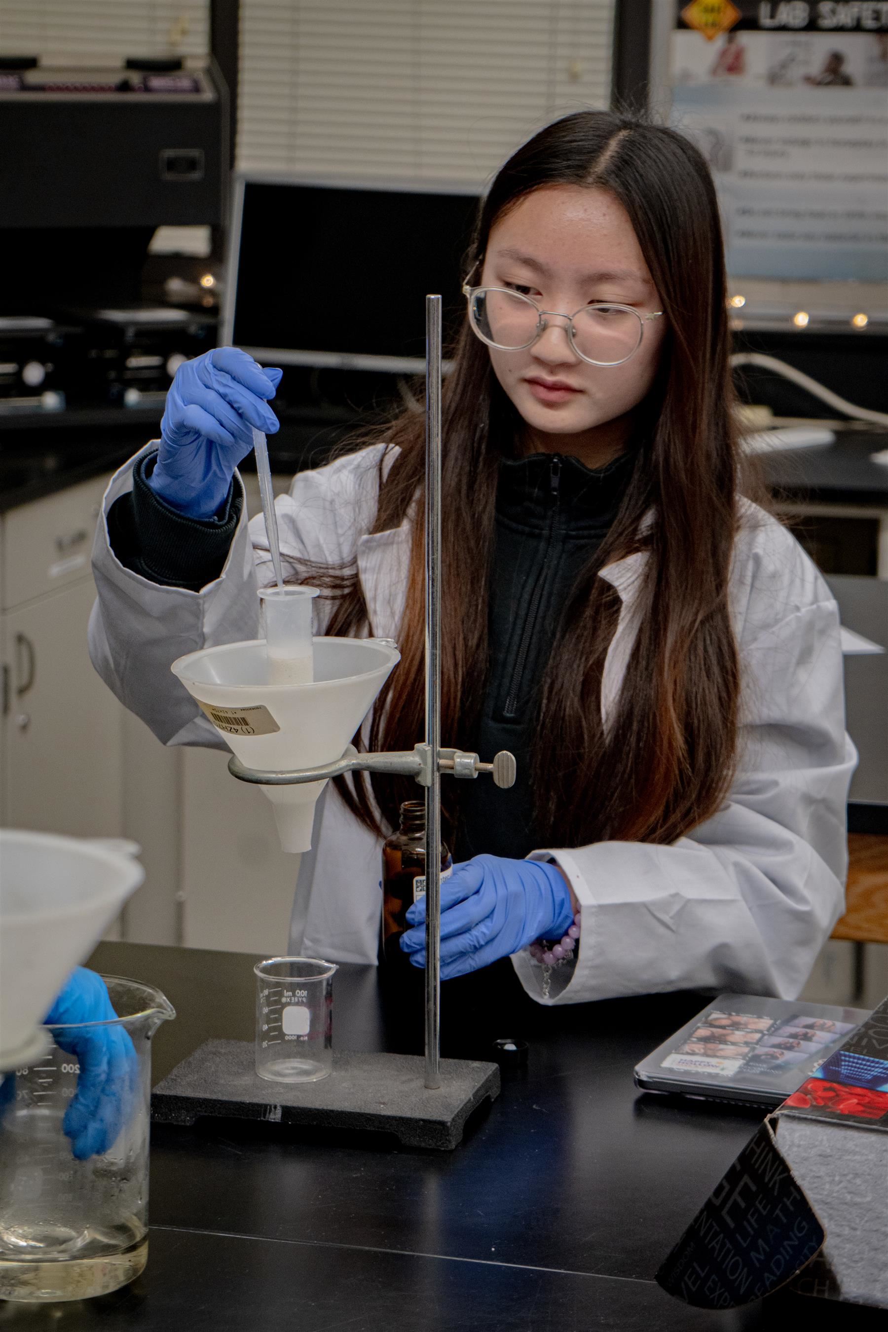 A young student wearing round glasses and blue protective gloves is focused intently on a chemistry experiment in a laborator